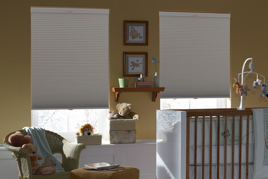 Cellular shades are a nifty option for energy-efficient and stylish nursery window treatments. Blackout gray cellular shades in two windows. Displayed inside a baby room.